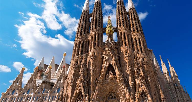 Vista hacia arriba de las agujas que se elevan de la Sagrada Familia contra un cielo azul brillante con nubes