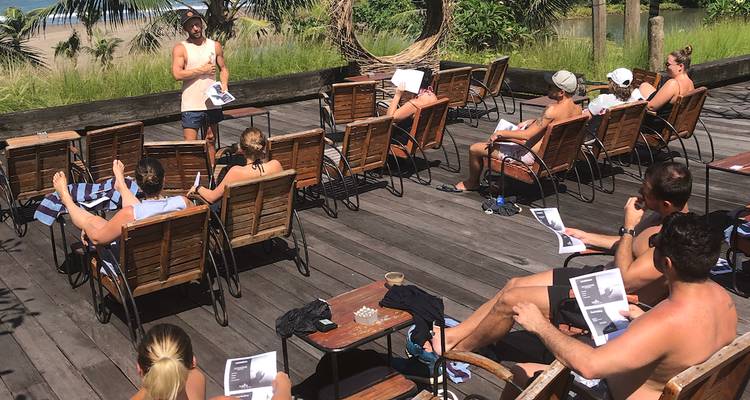Sesión de aula al aire libre con viajeros vestidos de manera casual sentados en tumbonas de madera escuchando a un instructor