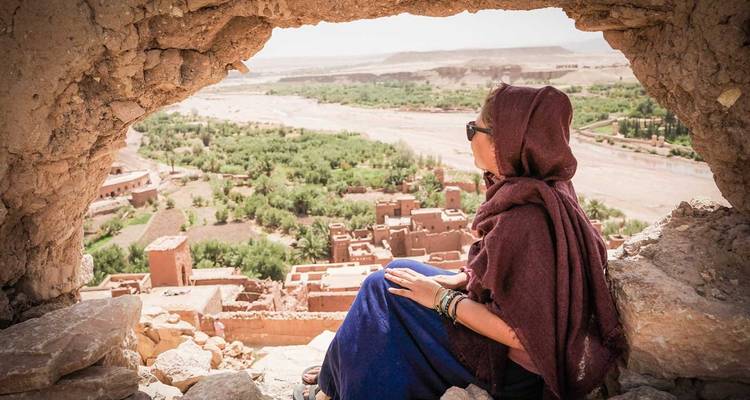 Traveler in a headscarf sits inside a mud-brick window opening overlooking the verdant valley and ruins of Ait Benhaddou