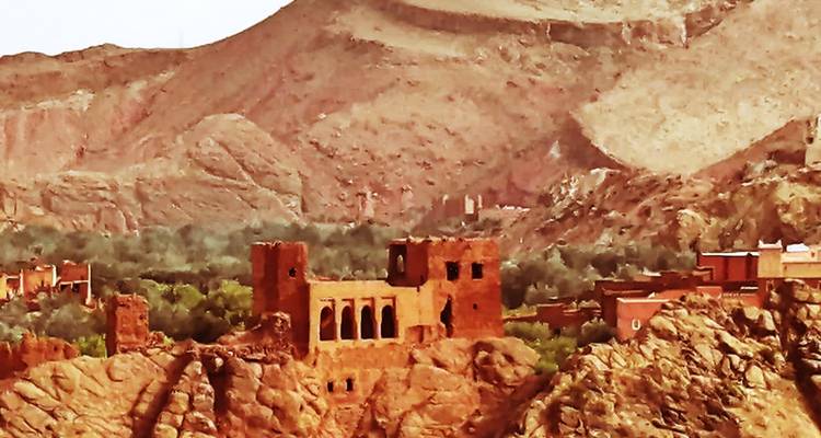 Ruined red-stone kasbah perched on rocky outcrop overlooking green valley and arid mountains