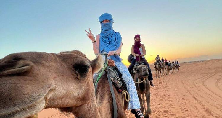 Close-up of smiling traveler on camel giving a peace sign while a caravan winds across sandy desert at sunset