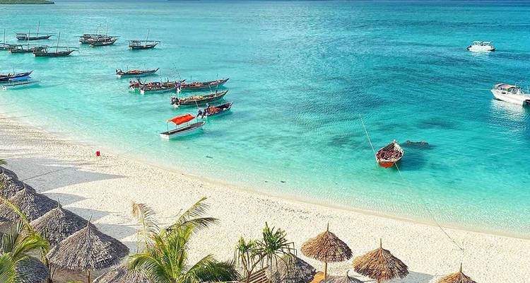 Une plage avec des bateaux dans des eaux turquoise et des parasols de chaume.