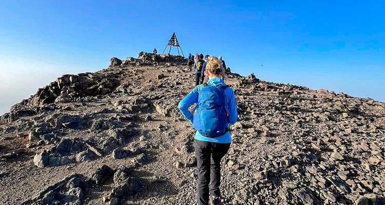 Les trekkers s'approchent de la pyramide de sommet en métal du Mont Toubkal sous un ciel bleu éclatant.