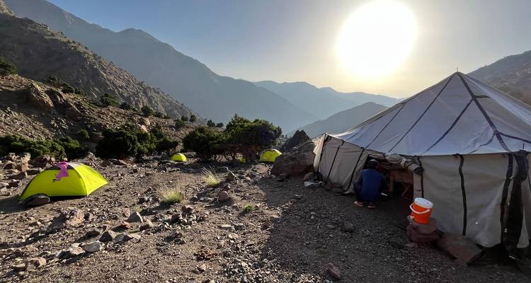Un petit camping sous tentes se situe sur un flanc montagneux rocheux tandis que le soleil se couche derrière des crêtes brumeuses.