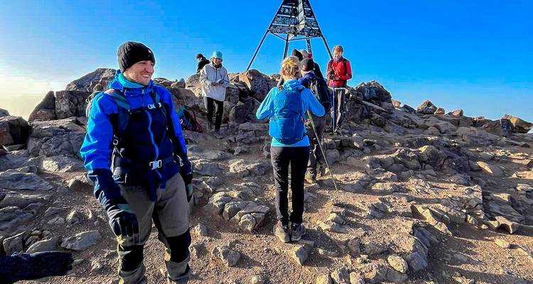 Les grimpeurs célèbrent au sommet rocheux du Toubkal à côté de la structure métallique emblématique sous un ciel dégagé.