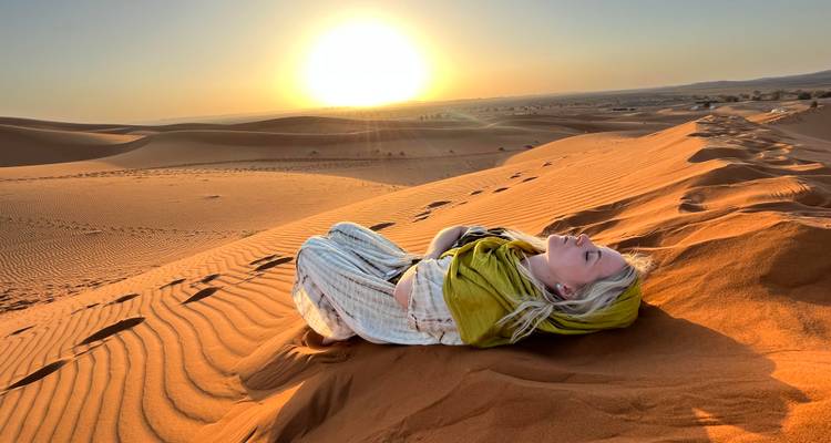 Mujer envuelta en una bufanda se reclina sobre una duna de arena dorada mientras el sol se pone sobre el desierto ondulante.