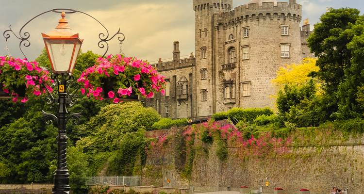 Elegante stenen muren en torens van Kilkenny Castle rijzen boven bloeiende roze bloemen en een lantaarnpaal uit boven het gouden uur.
