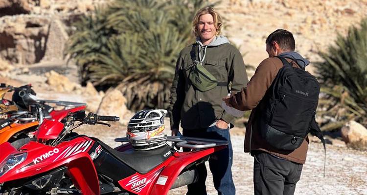 Two individuals beside a quad bike in a rocky, desert area.
