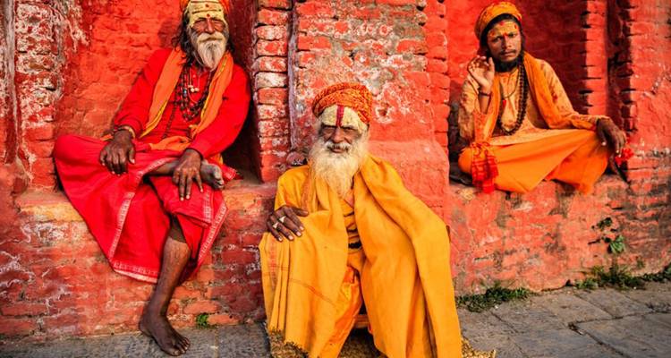 Three colorfully robed sadhus relax against a bright red temple wall, one gesturing in blessing.