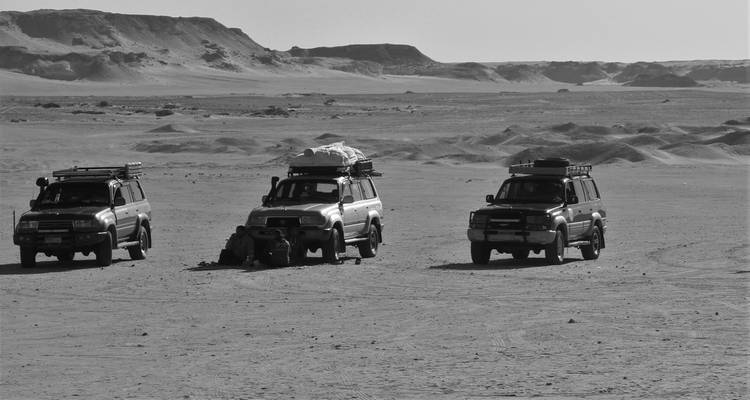 Image en noir et blanc de trois véhicules tout-terrain garés sur de vastes sables du désert avec des plateaux lointains.