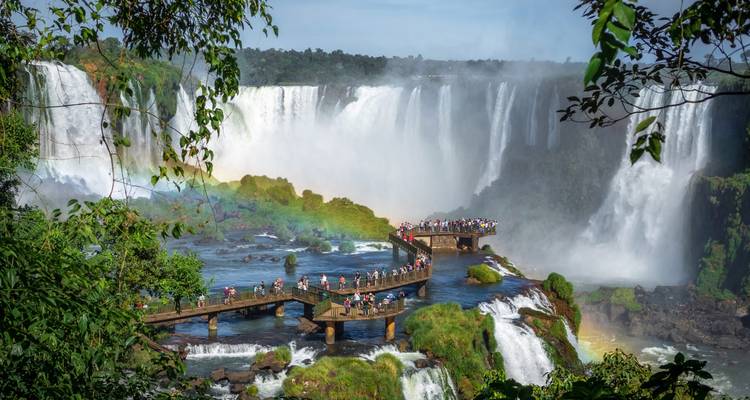 Panoramic view of Iguazu Falls with a rainbow arcing over a long viewing boardwalk filled with visitors