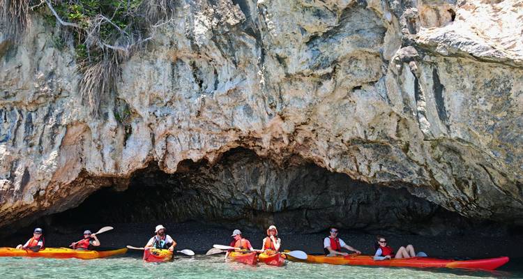Una fila de kayakistas en botes de colores brillantes explora una cueva marina rocosa a lo largo de una costa turquesa cristalina.