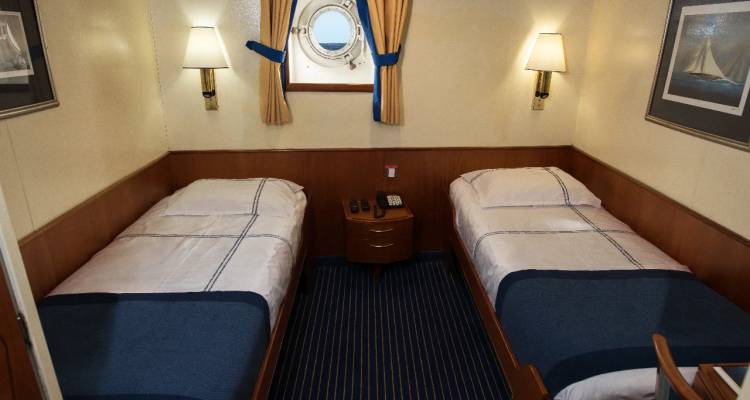 Compact twin-bed cruise cabin with porthole window, striped carpet and wall-mounted lamps.