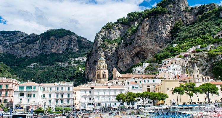 Italiaanse kustplaats met stenen klokkentoren tegen hoge groene kliffen onder heldere wolken.