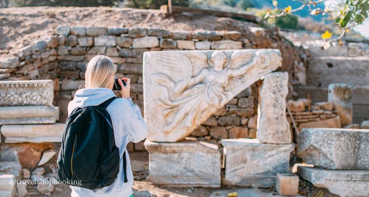 Female traveller photographs an angel relief among ancient marble ruins; watermark visible bottom left.