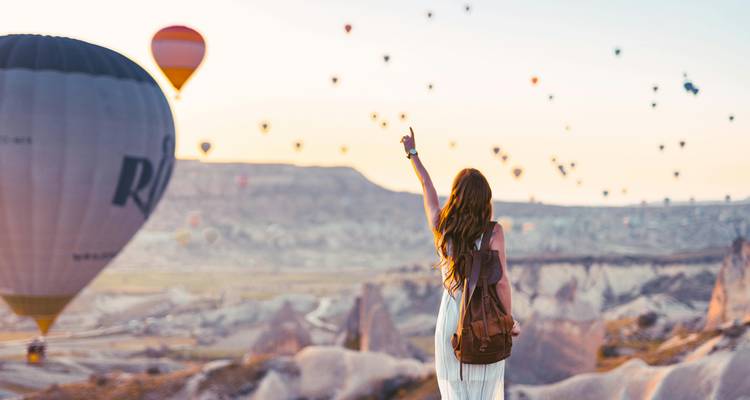 Back-view of a woman pointing toward dozens of hot-air balloons floating over Cappadocia's fairy-chimney landscape at sunrise.
