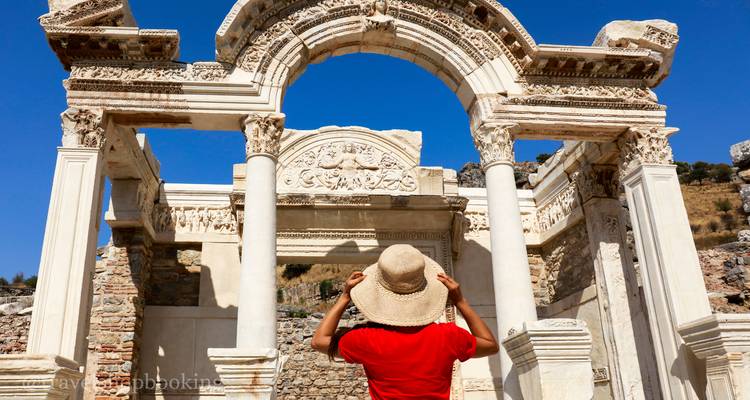 A woman in a red shirt and straw hat admires the ornate marble arch of the Temple of Hadrian in Ephesus.