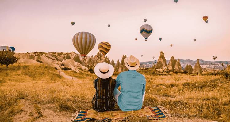 Una pareja con sombreros se sienta en una alfombra observando docenas de globos aerostáticos flotar sobre Capadocia al amanecer.