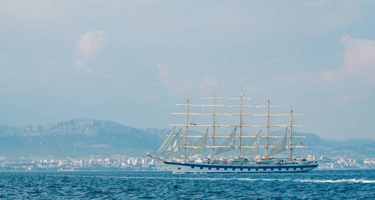 Grand voilier sur la mer avec ville et montagne en arrière-plan.