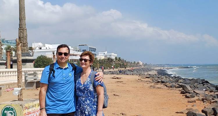 Couple standing in front of a rocky beach lined with buildings in the background.