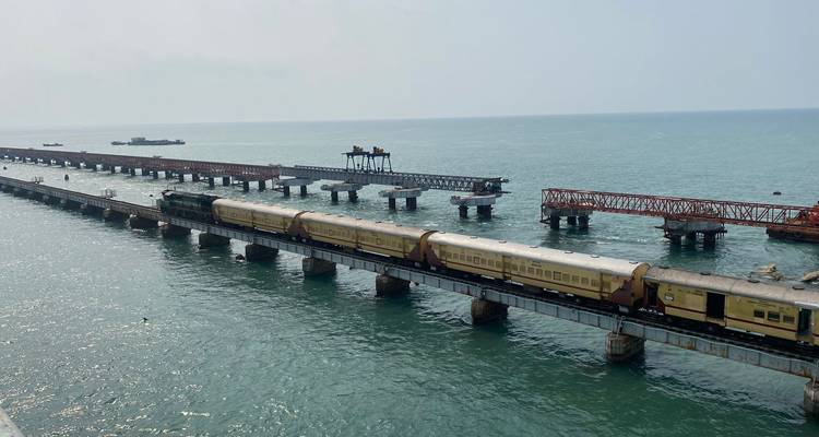 A train crossing a long bridge over the sea with distant industrial structures.