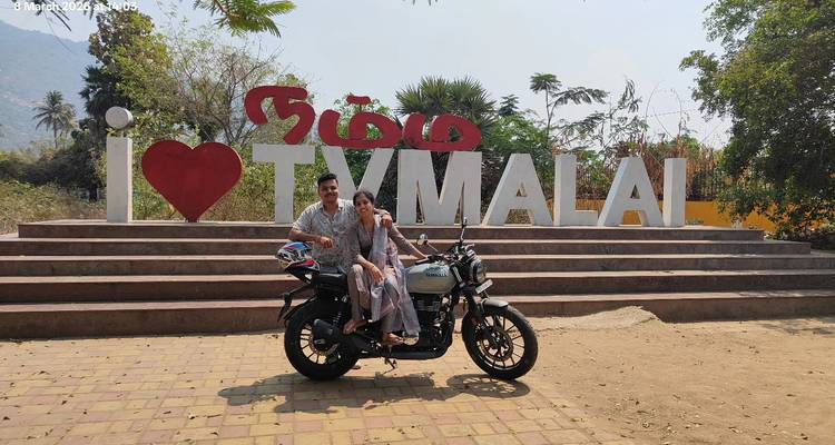 Young couple sitting on a motorbike in front of an 'I ❤️ TYMALAI' sign on a sunny day.