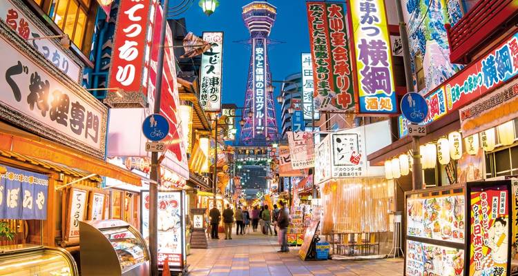 Belebte Straße in Osaka mit leuchtenden Werbeanzeigen und dem Tsutenkaku-Turm im Hintergrund.
