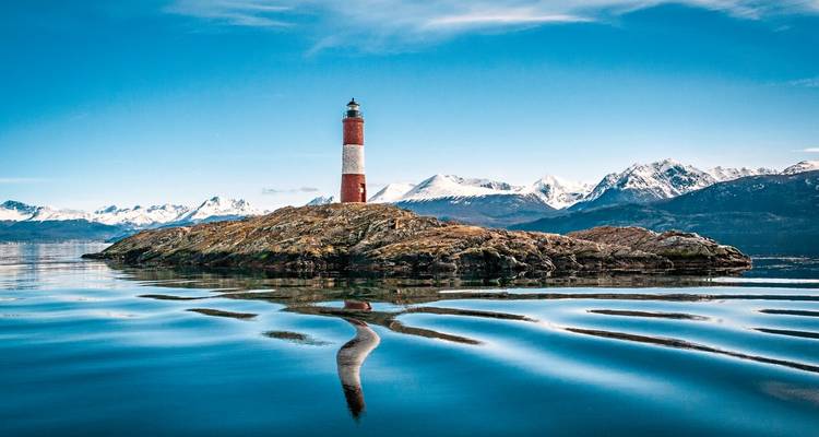 Rot-weißer Leuchtturm auf einer felsigen Insel, mit schneebedeckten Bergen im Hintergrund.