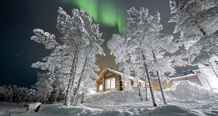 Verschneite Landschaft mit Nordlichtern darüber und einer Holzhütte.