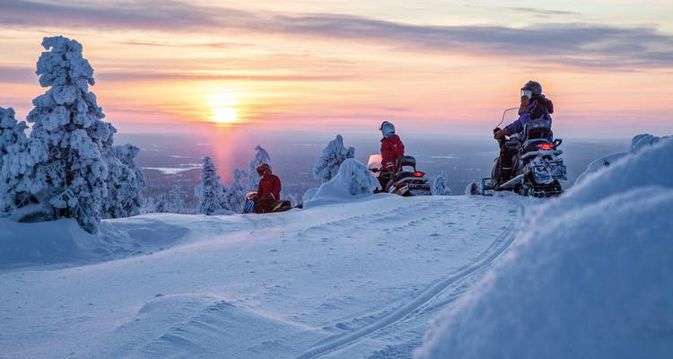 Gruppe fährt Schneemobile in einer verschneiten Landschaft während des Sonnenuntergangs.