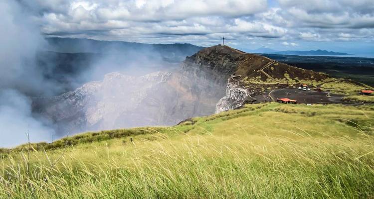 Plateau herbeux surplombant le cratère fumant du volcan Masaya sous un ciel nuageux dramatique