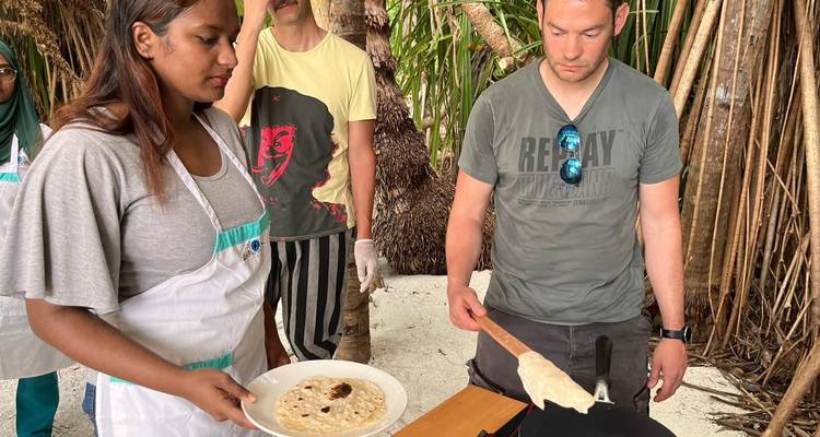 Los viajeros participan en una clase de cocina maldiviana al aire libre, preparando pan plano sobre una plancha caliente entre palmeras.