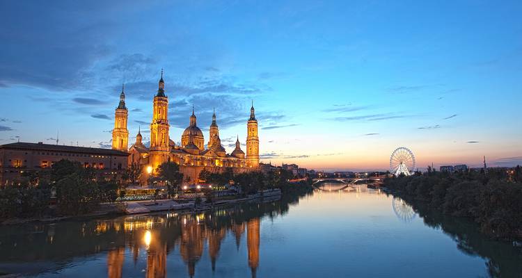 Basílica de Nuestra Señora del Pilar brillando junto al río Ebro en la hora azul con noria a la distancia.