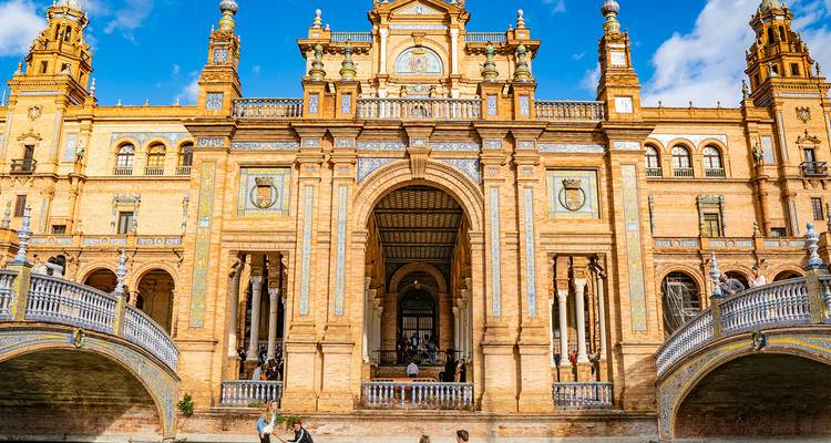 Gran fachada de la Plaza de España con arcos ornamentados, azulejería y visitantes cruzando puentes.