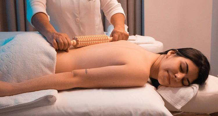 Een spa-therapeut gebruikt een houten roller om de rug van een ontspannen vrouw die op een behandeltafel ligt te masseren.