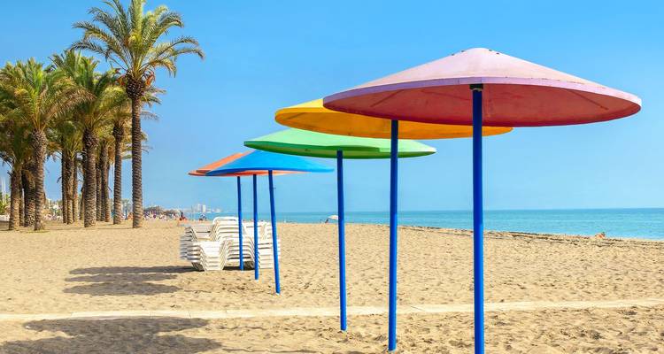 Farbenfrohe Sonnenschirme aufgereiht an einem sandigen Mittelmeer-Strand neben Palmen und ruhigem türkisfarbenem Wasser