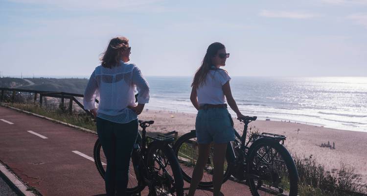 Dos mujeres se detienen con sus bicicletas en un carril para ciclistas junto al mar, mirando hacia una playa que brilla bajo el sol.