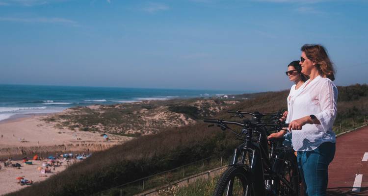Los ciclistas están de pie junto a sus bicicletas en un camino para bicicletas en un acantilado con vistas a una playa de arena abajo.