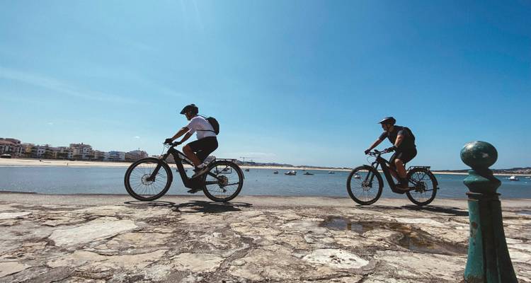 Dos ciclistas pedalean por un paseo marítimo junto a aguas azules tranquilas bajo un cielo despejado.