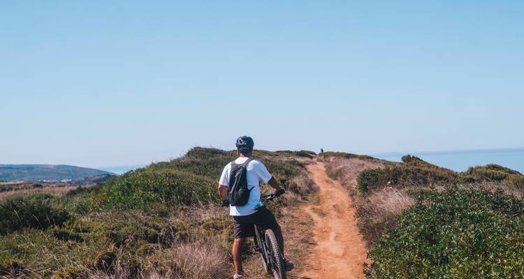 Un ciclista está de pie con su bici en un estrecho sendero de cresta de tierra que domina la maleza y el mar distante.