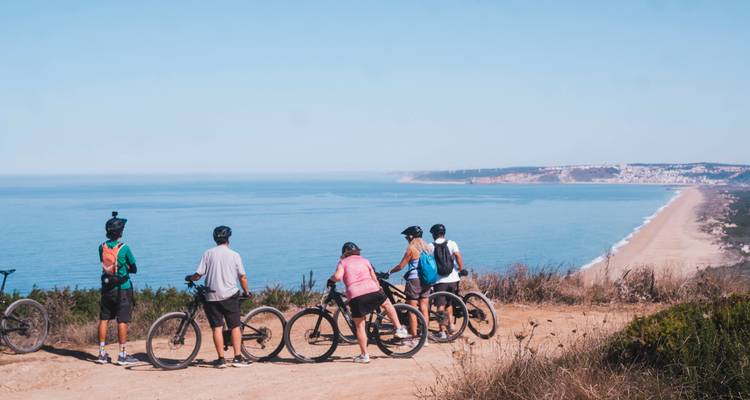 Un grupo de ciclistas de montaña se reúne en un mirador acantilado costero muy por encima de una amplia playa de arena.
