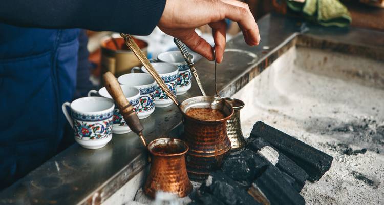 Preparar café turco en una cezve de cobre sobre arena caliente con tazas ornamentadas cerca.