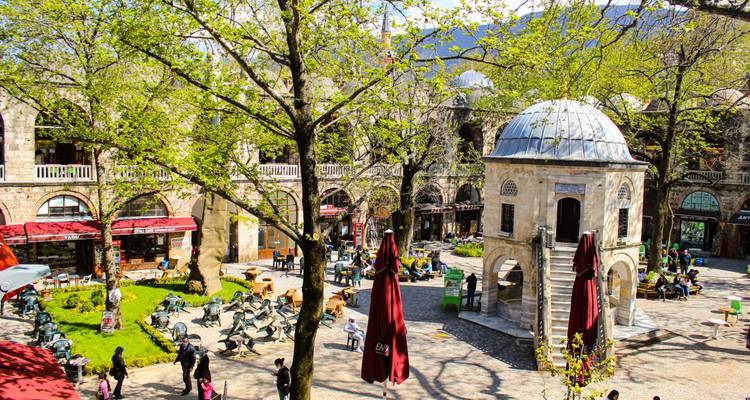 Patio animado de Koza Han con follaje primaveral, cafés al aire libre y gente relajándose.