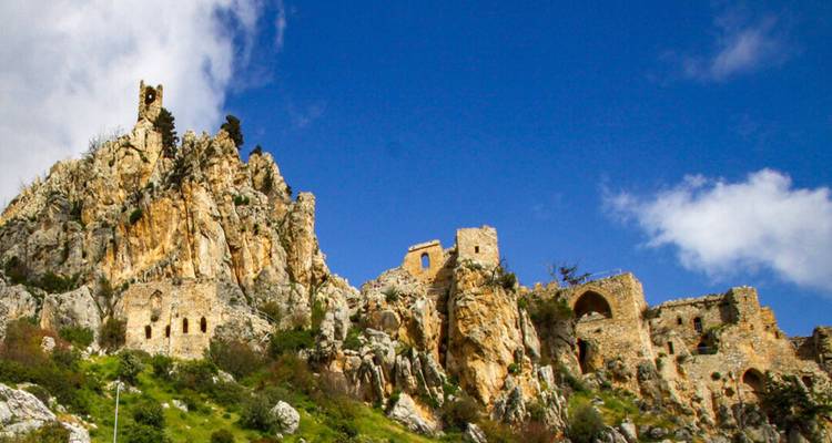 Ruined stone walls and towers of St. Hilarion Castle rising dramatically on a jagged rocky peak against a bright blue sky.