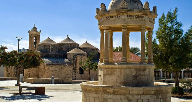 Historic stone fountain and domed Byzantine church set in a sunny plaza with trees.