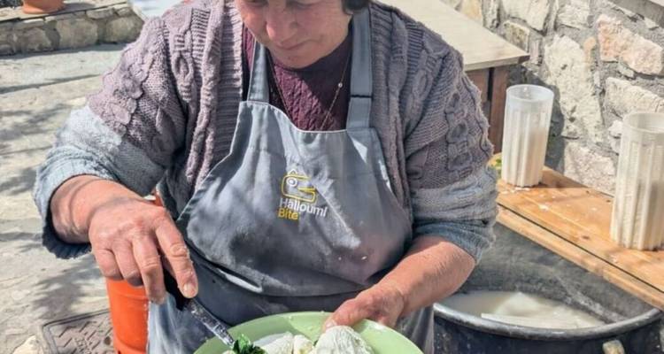 Local woman in an apron preparing fresh halloumi cheese outdoors beside stone walls.