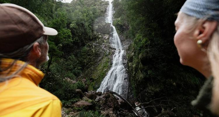 Two hikers admire a tall cascading waterfall deep in a forested ravine.