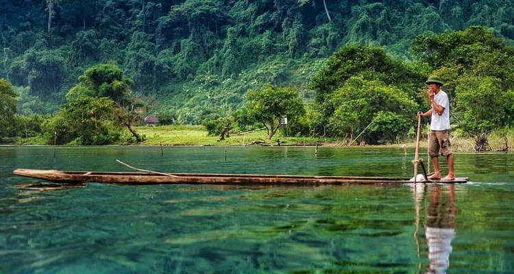 Homme sur un petit bateau ramant sur une rivière aux eaux vertes entouré par la nature.