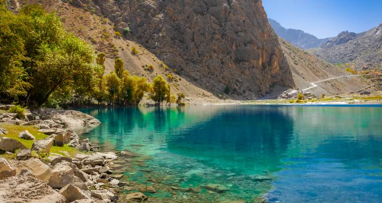 Turquoise alpine lake mirrors golden trees and towering rocky walls in the Fann Mountains.