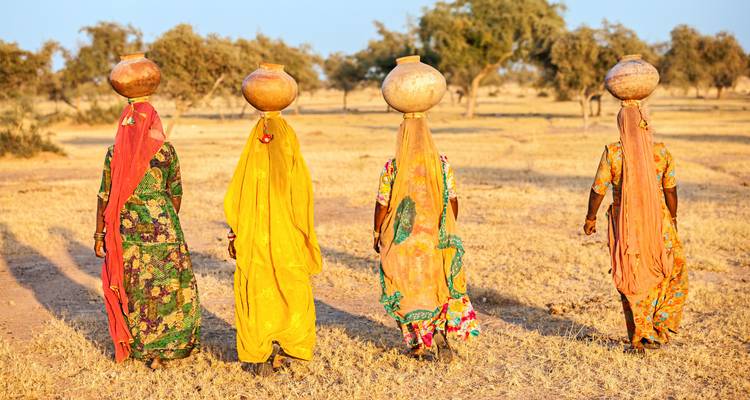 Women carrying pots on their heads walking across a dry field.